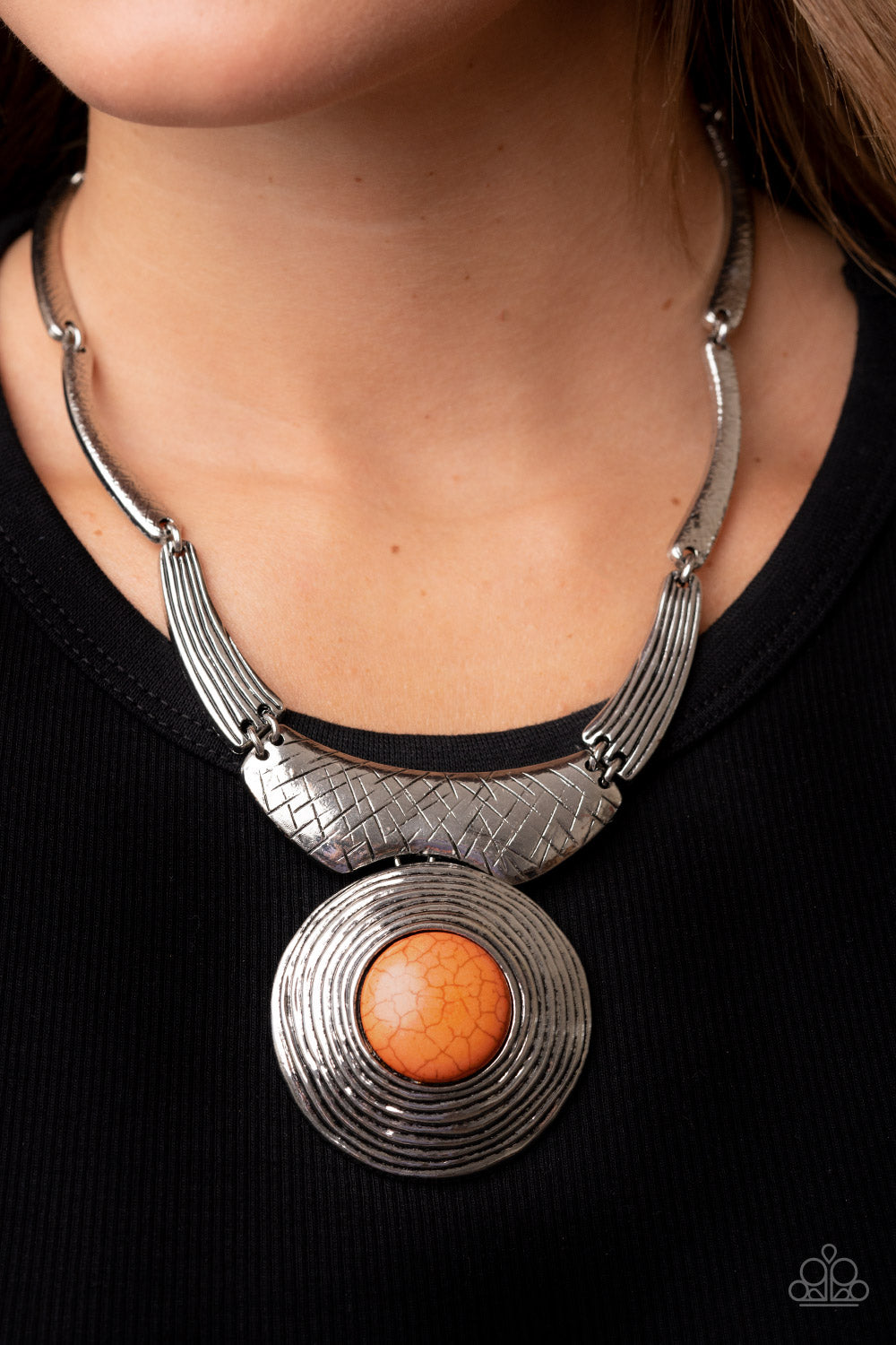 Featuring hammered, scratched, and linear patterns, an antiqued assortment of gently curving silver frames boldly links below the collar. Dotted with an oversized orange stone center, a rustic silver frame radiating with circular texture swings from the bottom for a dramatically earthy flair. Features an adjustable clasp closure.