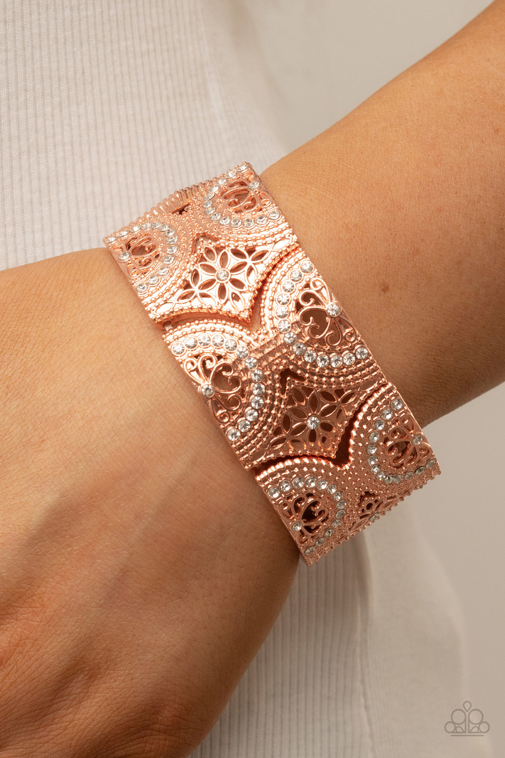 Dotted with sections of glassy white rhinestones, an immaculate display of studded wheel and floral shiny copper patterns combines into shimmering frames along stretchy bands around the wrist for a regal flair.