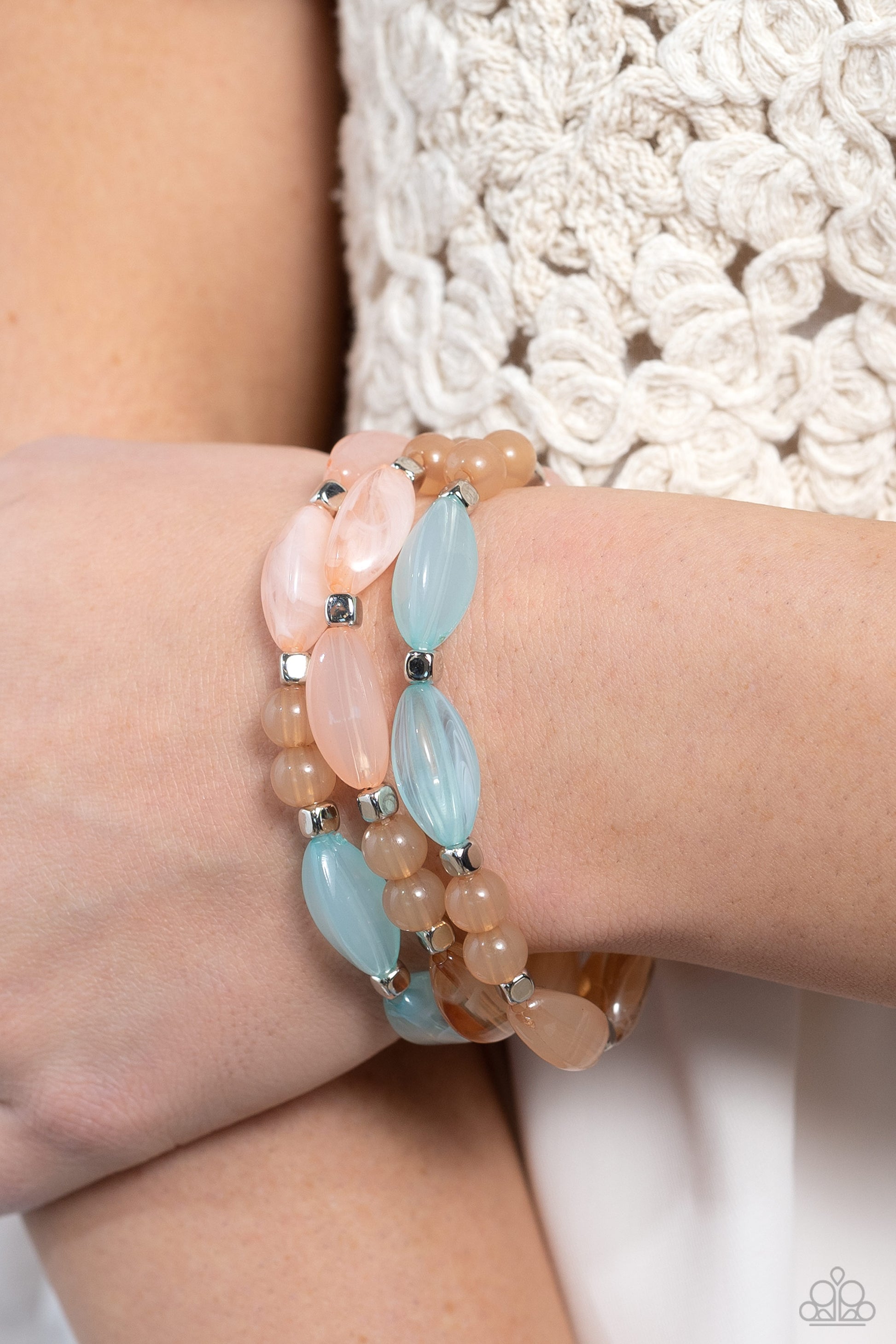 Shiny silver cubed accents, cloudy, milky, light blue, light pink, and peach oval beads and milky peach beads wrap around the wrist on stretchy bands, creating colorful layers.